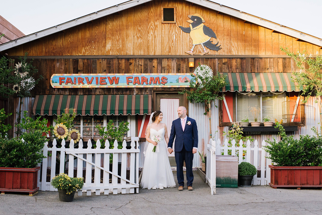 A wedding couple stands holding hands in front of a store front.