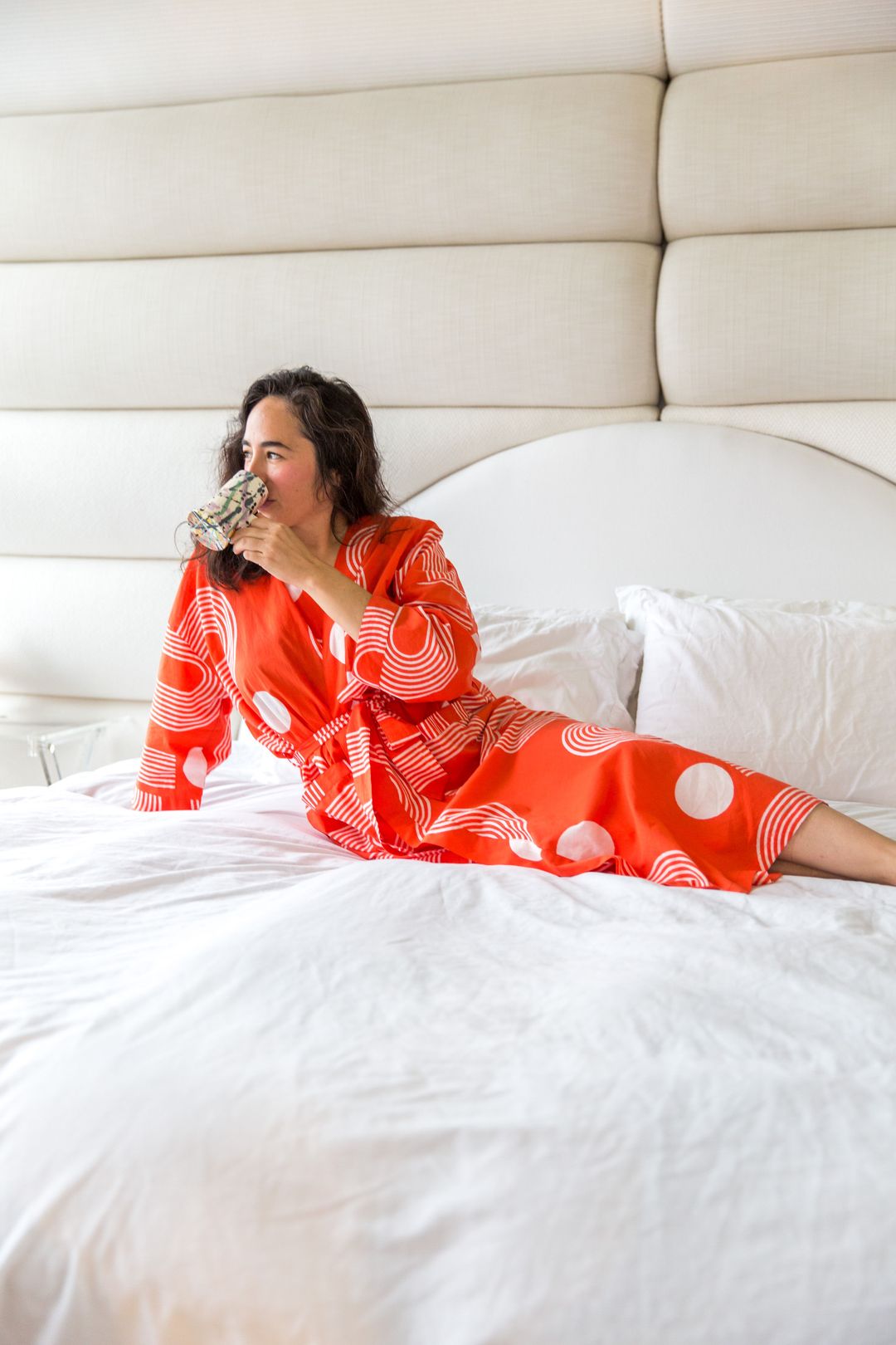 A woman lounges on a bed while sipping a drink and wearing a robe.