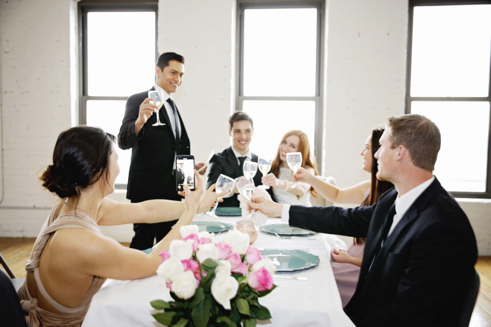 People at a wedding clinking glasses and recording on a phone.