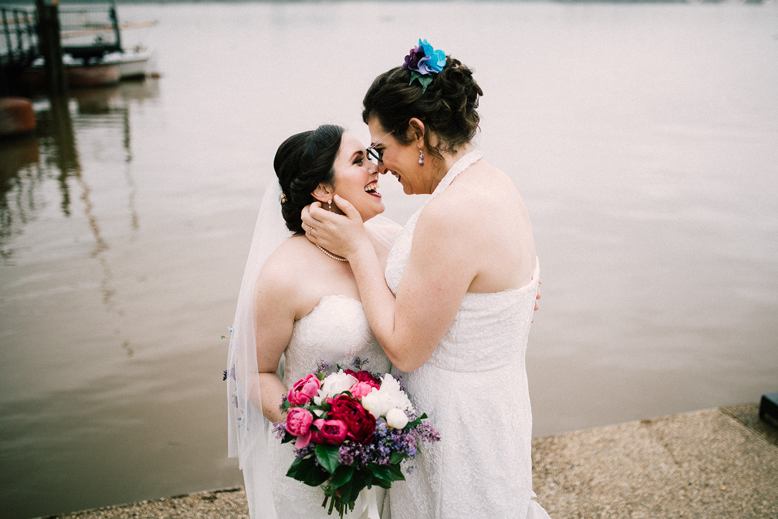 Two brides smile and hug at the shoreline.