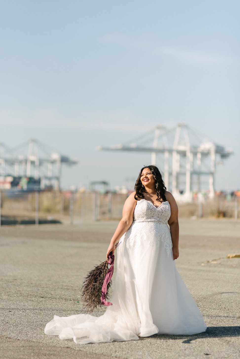 A woman stands in front of cranes smiling and wearing the Maggie Sottero dress style 'Vanessa.'