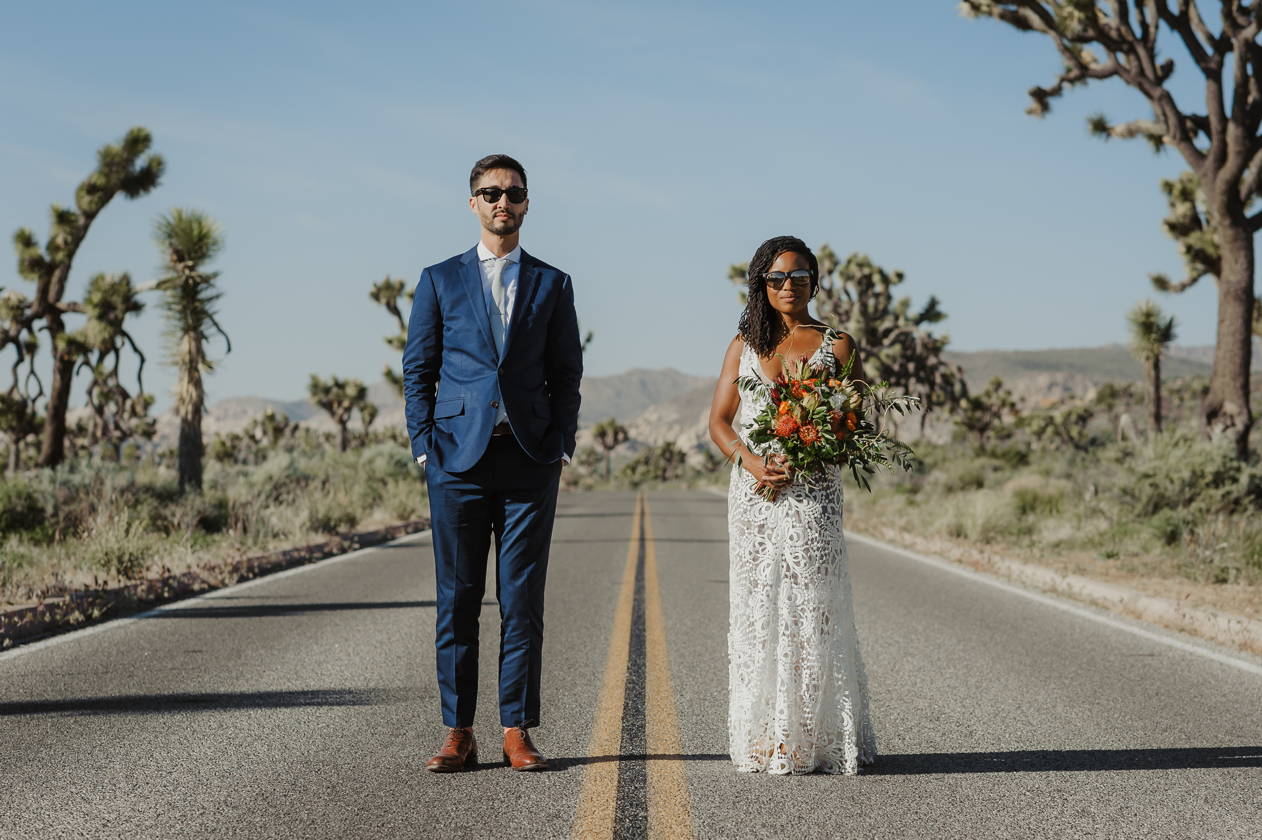 Groom in a blue suit and bride in a lace dress stand in the middle of a deserted highway in Joshua Tree