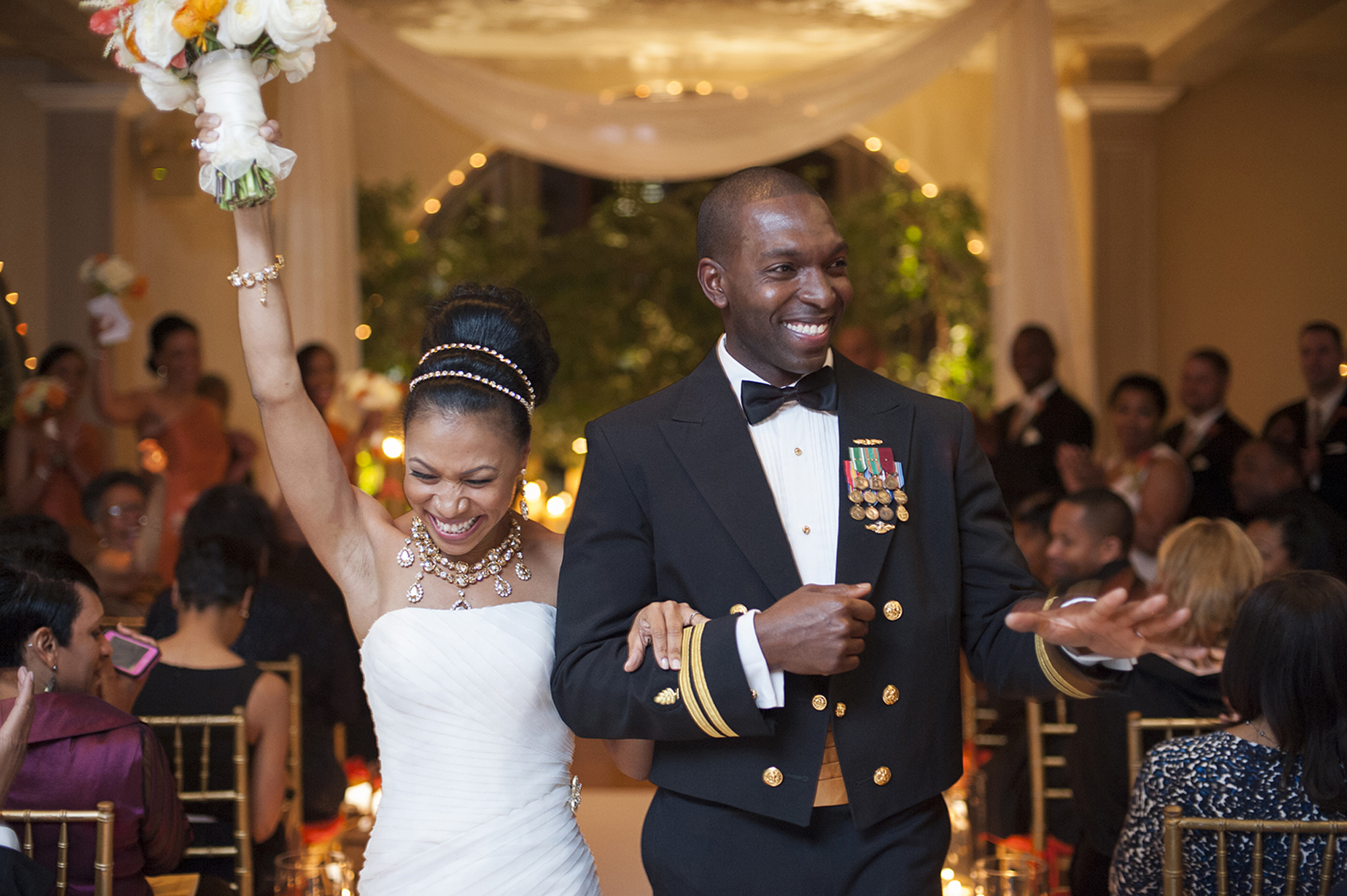 Black couple grinning as they walk down the aisle. Bride is raising up her bouquet and groom is wearing military dress.