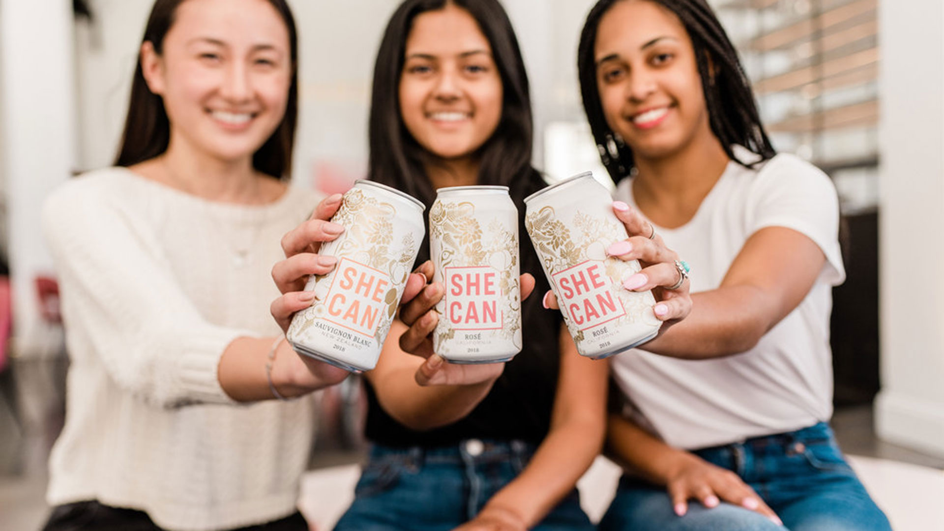 three womxn holding up "SHE CAN" wine cans