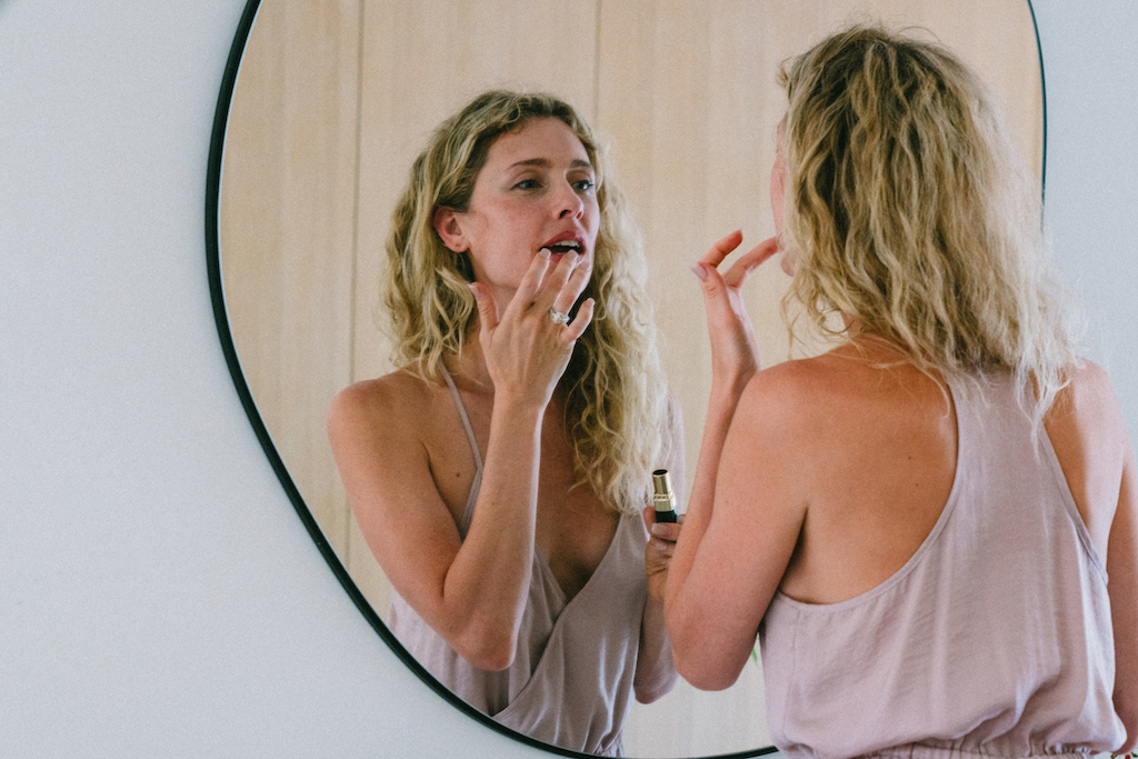 Woman looks into mirror, applying lipstick. You can see her engagement ring on her hand.