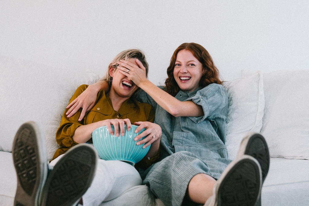 Two women sit on a couch with a bowl of popcorn. The woman on the right puts her hand over the other's eyes, and you can see her engagement ring.