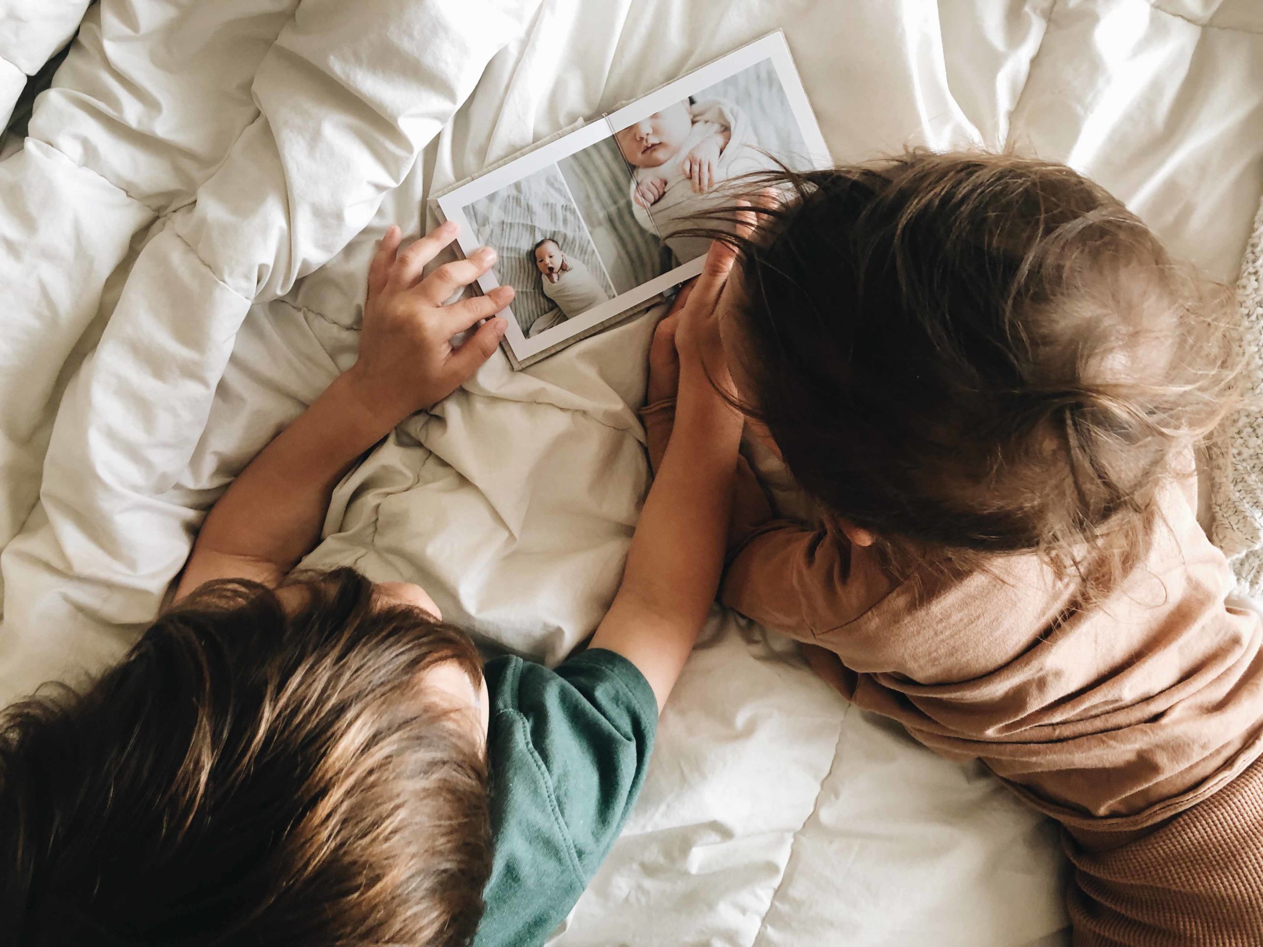 overhead shot of two small toddlers lying on a white comforter and looking at an Albums Remembered Little Hugs albums that is showing baby photos