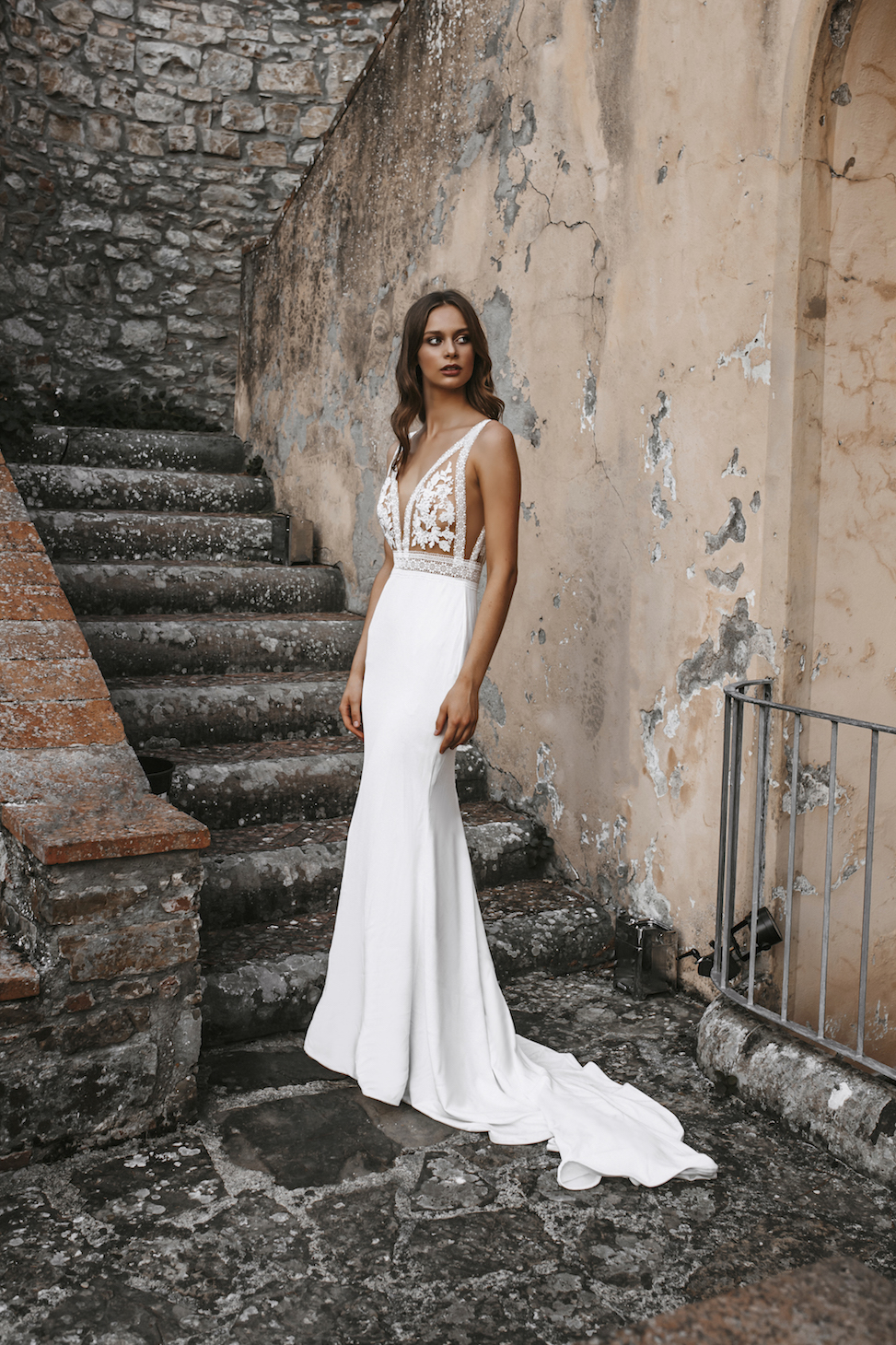Woman standing in front of run down stone stairs in white wedding dress with sheer and floral top