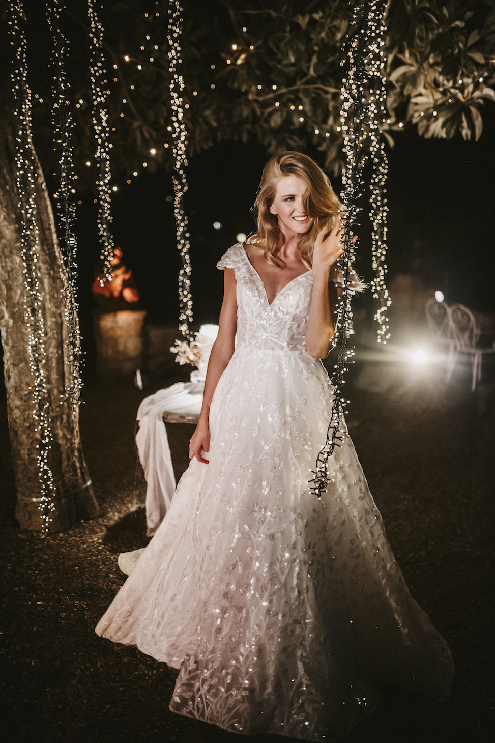 Woman outside among string lights, smiling, wearing a shimmery short sleeved white wedding dress from Allure Bridal