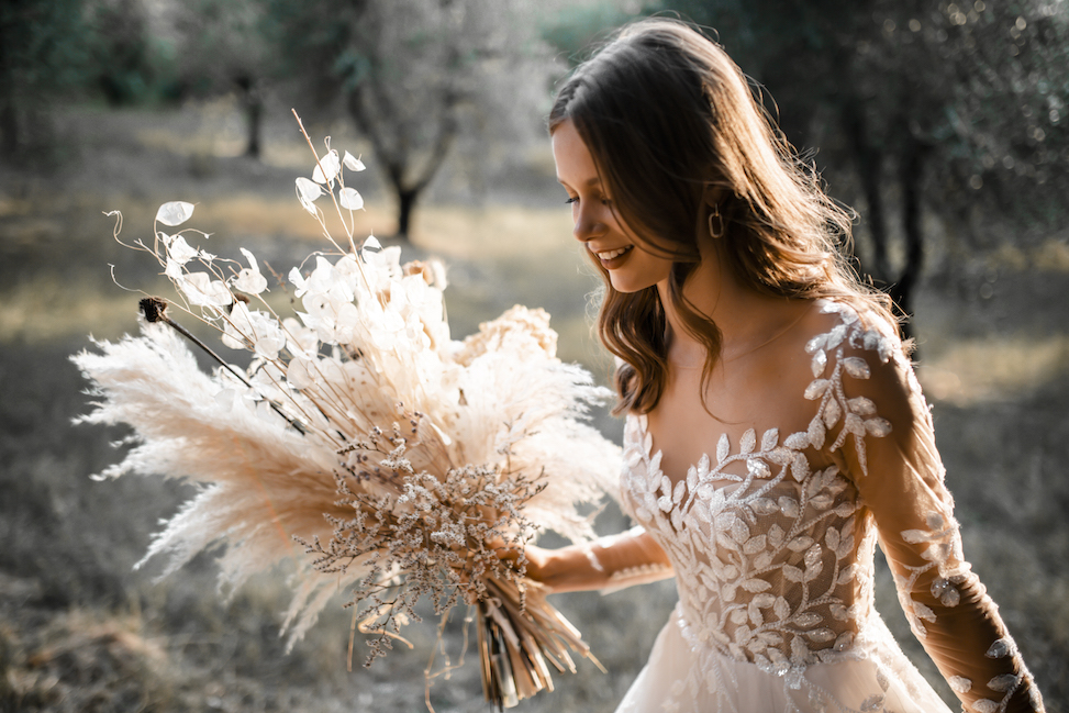 Woman in long sleeved, floral, white wedding gown holding dried florals, smiling, looking at the ground