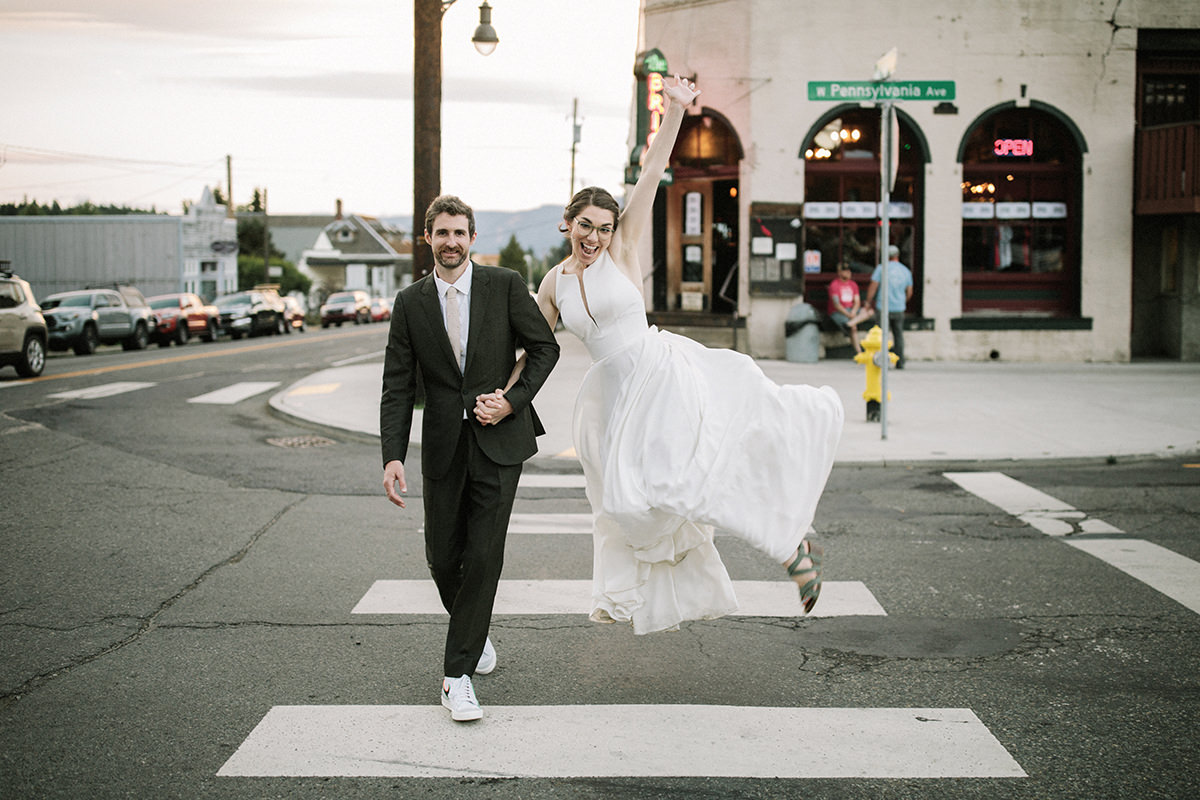 Groom and bride, holding hands, crossing the street, bride is jumping for joy.